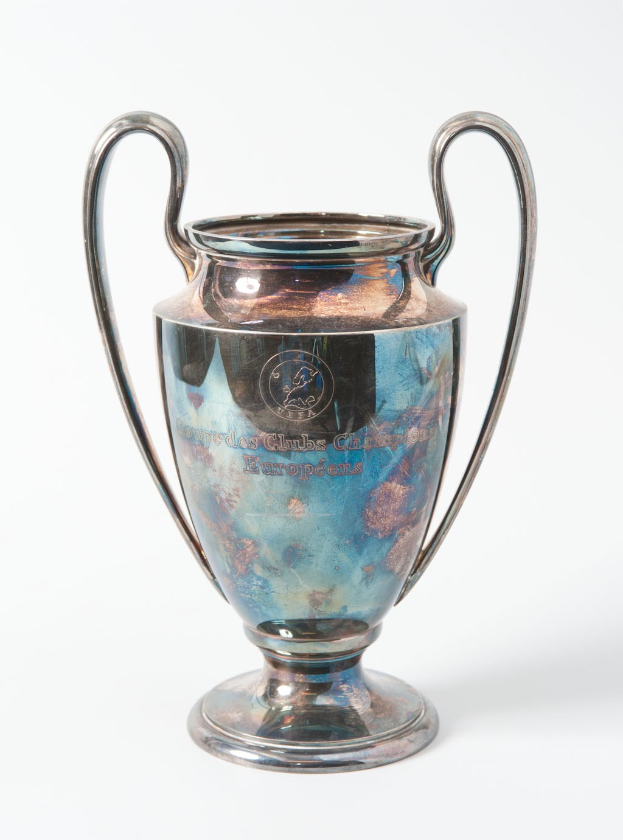 Der UEFA Champions League-Pokal, ein stahlerner Becher mit zwei Handles und einem blauen und silbernen Farbschema, der mit "UEFA Champions League" beschriftet ist und vor einem weißen Hintergrund steht.