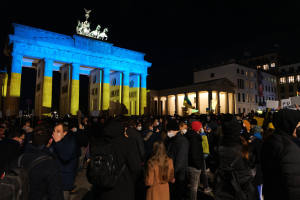 Eine Menschenmenge steht vor dem Brandenburger Tor in Berlin, Deutschland, viele tragen Mützen und haben Taschen dabei, einige halten Schilder, mit den Säulen und Statuen des Tors im Hintergrund, Gebäuden mit Fenstern im Hintergrund, unter einem dunklen Himmel.