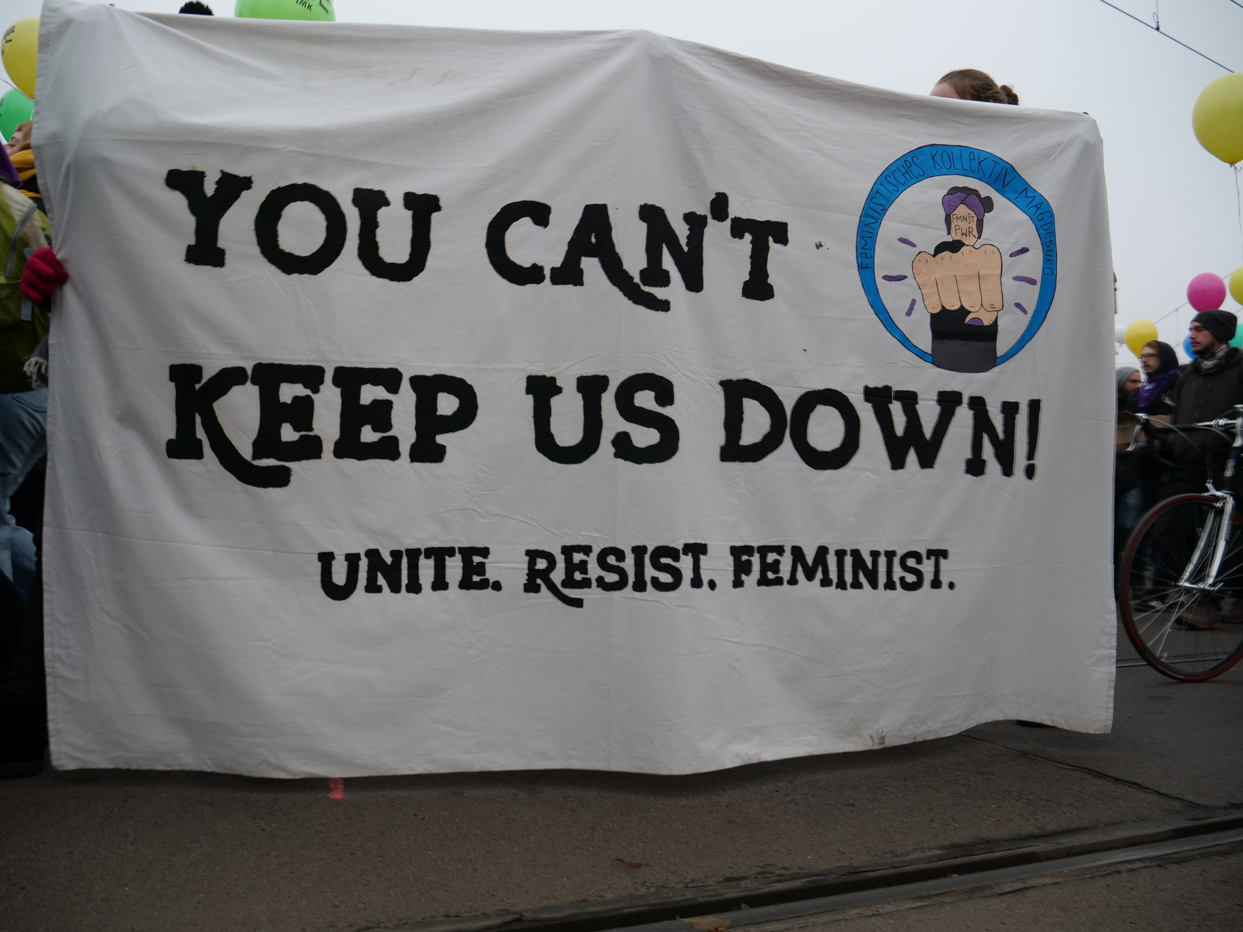 Eine Gruppe von Menschen steht auf der Straße und hält ein Banner in der Hand, auf dem "You Can't Keep Us Down, Unite Resist Feminist" steht, mit einem Fahrrad in der Nähe und Ballons, Drähten und einem klaren blauen Himmel im Hintergrund.