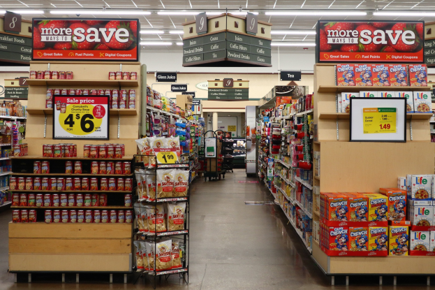 Ein Inneneinrichtungsbild eines Lebensmittelgeschäfts mit Regalen und Boden voller verschiedener Lebensmittel, beleuchtet von Deckenlampen und mit Informationsschildern mit Text und Zahlen; im Hintergrund ist eine Person zu sehen.