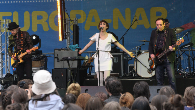 Eine Bühne mit vier Musikern: eine singende Frau in einem weißen Kleid in der Mitte, zwei Gitarristen links und rechts und eine Menge und Lautsprecher im Hintergrund.