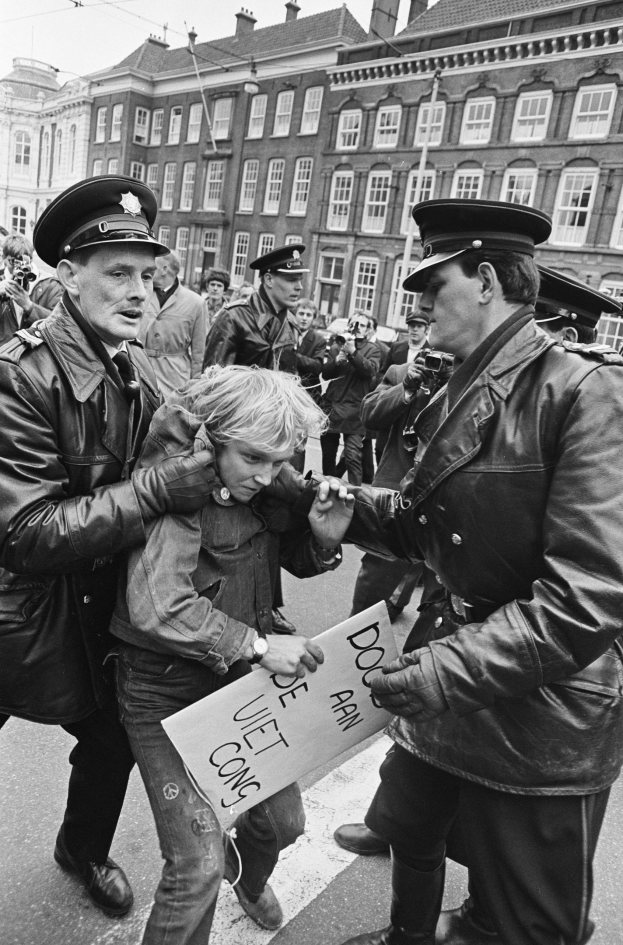 Schwarzes und weißes Bild einer Frau, die von zwei Polizisten auf einer Straße eskortiert wird, hält ein Blatt Papier und wird von einem Officer festgehalten, mit Menschen, Kameras, Gebäuden, Drähten und Himmel im Hintergrund.