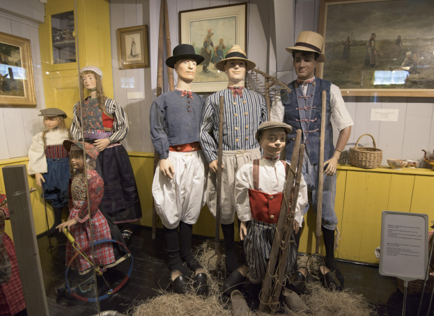 Eine Gruppe von Schaufensterpuppen in historischer Kleidung steht zusammen in einem Raum mit trockenem Gras auf dem Boden, einem Textschild, wandmontierten Foto-Rahmen, einem Schrank mit Gegenst├Ąnden und einem Korb und anderen Gegenst├Ąnden auf einer Plattform, wahrscheinlich im Nationalen Historischen Museum der Vereinigten Staaten.