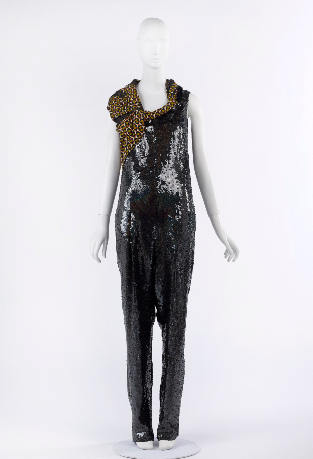 Ein Schaufensterpuppen-Mann in einem schwarzen und goldenen Pailletten-Jumpsuit mit einem Leoparden-Muster-Schal um den Hals, die Pailletten glitzern und das Licht reflektieren.