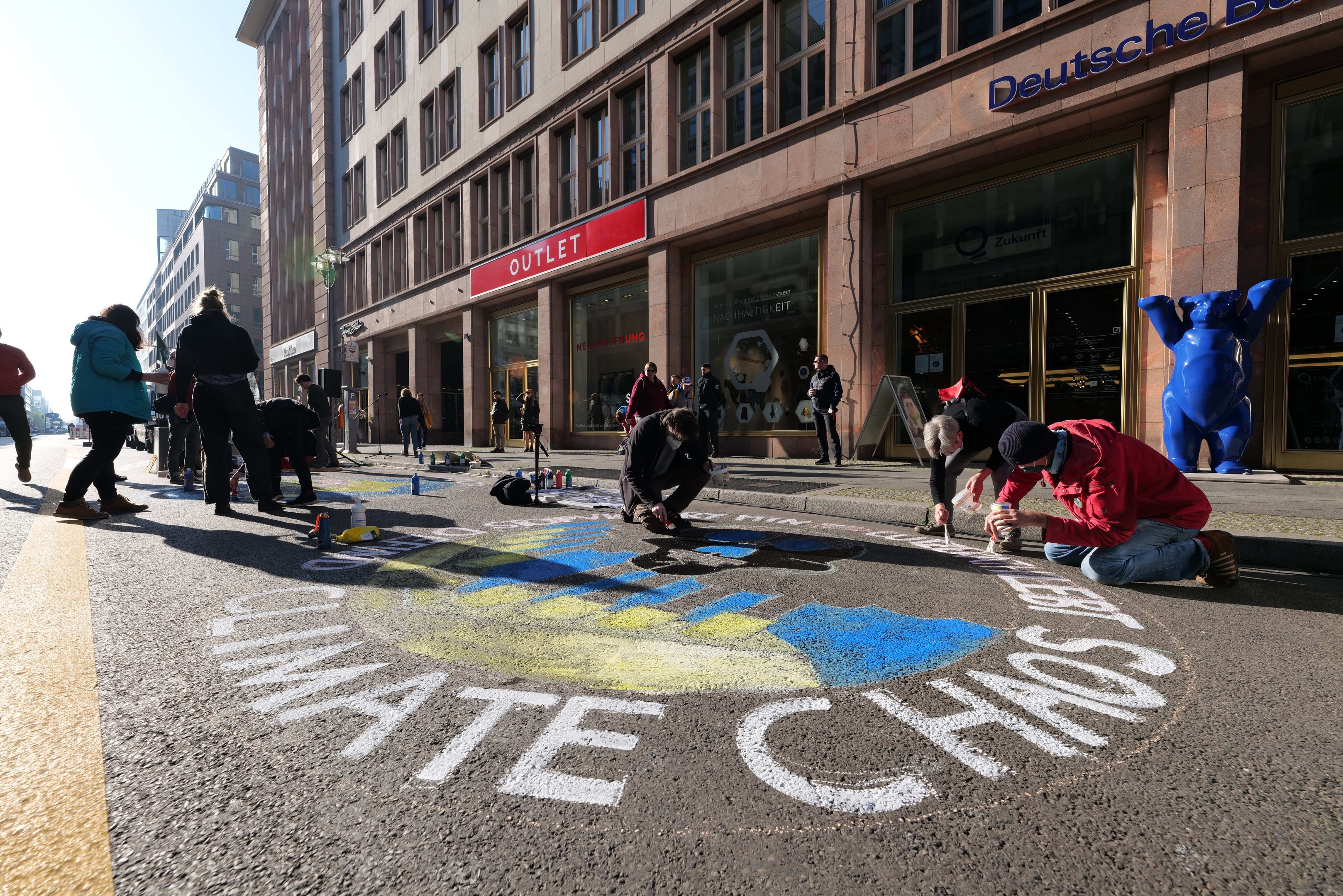 Menschen sitzen vor einem Gebäude während einer Klimaprotest in Berlin, umgeben von Flaschen und anderen Gegenständen, mit Bäumen und einem klaren blauen Himmel im Hintergrund.