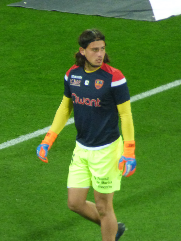 Ein Mann in einem blauen T-Shirt und gelben Shorts läuft auf einem Fußballfeld, die Arme ausgestreckt und mit entschlossener Miene, mit einem Tuch oben auf dem Bild.