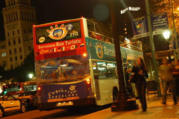 Fahrzeuge und ein Bus mit Fahrgästen fahren auf einer Straße, während Fußgänger auf dem benachbarten Gehweg gehen; Laternen, Banner, Bäume und Gebäude sind im Hintergrund sichtbar.