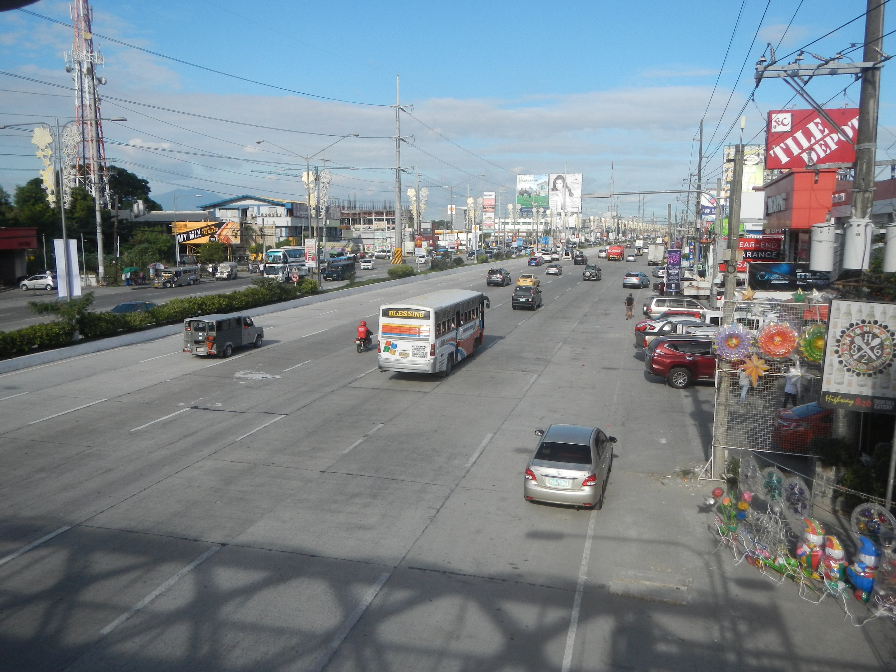 Eine belebte Stadtstraße mit verschiedenen Fahrzeugen, Strommasten, Gebäuden mit Namensschildern, Bäumen, Pflanzen und einem Turm unter einem bewölkten Himmel.