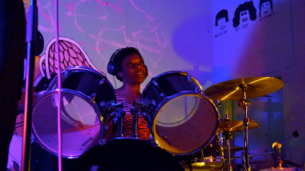 Eine Frau, die Musikinstrumente auf einer B├╝hne spielt, mit einem Mikrofonstativ und einer Person im Hintergrund.