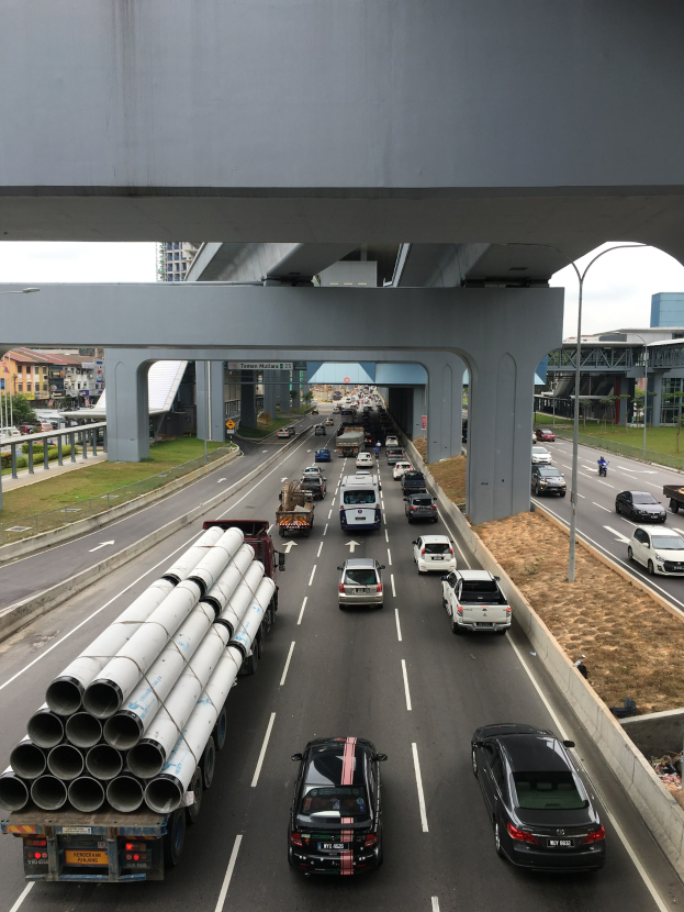 Eine stark befahrene Autobahn mit Schwerlastverkehr unter einer Brücke, umgeben von Gras, Bäumen, Gebäuden und Strommasten.