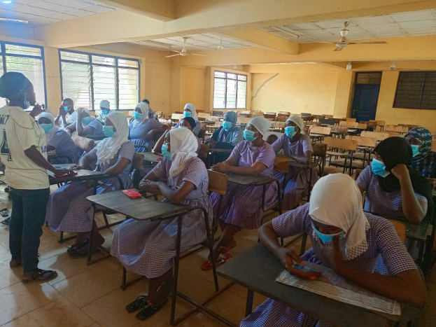Eine Klasse in Nigeria mit Schülerinnen und Schülern, die Gesichtsmasken tragen und auf Bänken sitzen oder stehen, mit Büchern und Gegenständen auf Tischen, Fenstern, einer Tür und Deckenlampen und -ventilatoren im Hintergrund.
