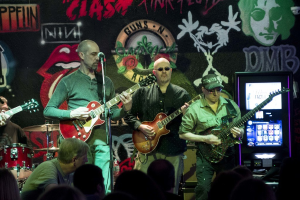 Drei Männer spielen Gitarren auf einer Bühne mit einer Menge darunter und Graffiti an der Wand im Hintergrund.