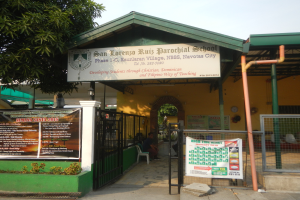 Der Eingang der San Lorenzo Ruiz Pfarrschule hat ein Schild mit Text, einen Metallzaun, Menschen auf Stühlen unter einem Dach, Pfosten, ein Schild, Pflanzen, einen Baum und einen bewölkten Himmel.
