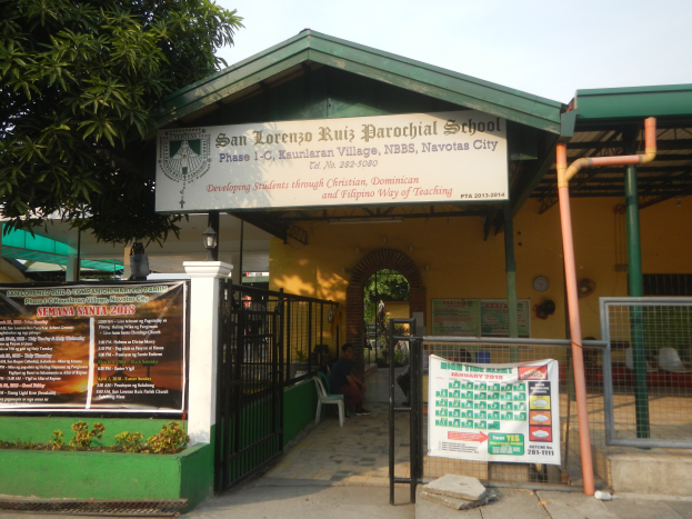 Der Eingang der San Lorenzo Ruiz Pfarrschule hat ein Schild mit Text, einen Metallzaun, Menschen auf Stühlen unter einem Dach, Pfosten, ein Schild, Pflanzen, einen Baum und einen bewölkten Himmel.