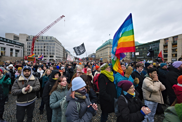 Große Gruppe von Menschen bei einer LGBTQ+-Rechtsdemo in Berlin, die Fahnen und Plakate schwenkt, mit Gebäuden, einem Kran und einem bewölkten Himmel im Hintergrund.
