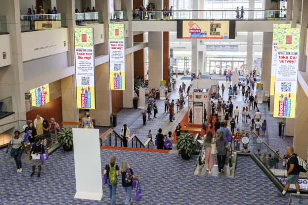 Eine große Gruppe von Menschen geht durch ein großes Gebäude mit Bannern, Topfpflanzen, Rolltreppen, Geländern, Säulen, Texttafeln und Glastüren, einige tragen Taschen, wahrscheinlich nehmen sie an der Nationalen Buchmesse teil.