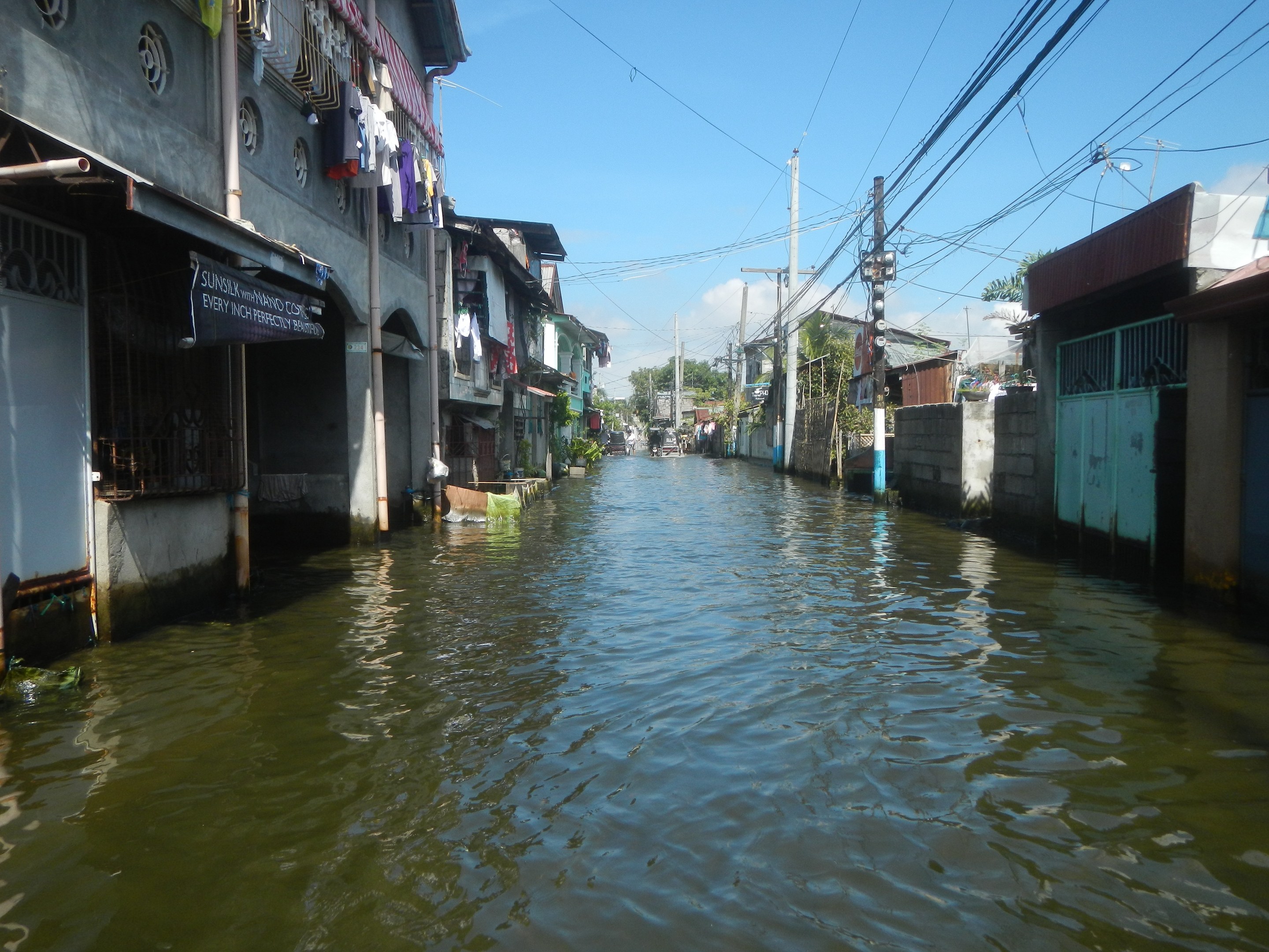 Flutstraße in der Stadtmitte mit Wasser bedeckt, Gebäuden auf beiden Seiten, Strommasten mit Drähten, zum Trocknen aufgehängter Wäsche, Bäumen und einem klaren blauen Himmel im Hintergrund.
