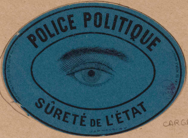 Ein brauner Karton mit einem blauen Aufkleber, auf dem "Police Politique S├╝reté de L'Etat" steht und ein Auge in der Mitte zeigt.