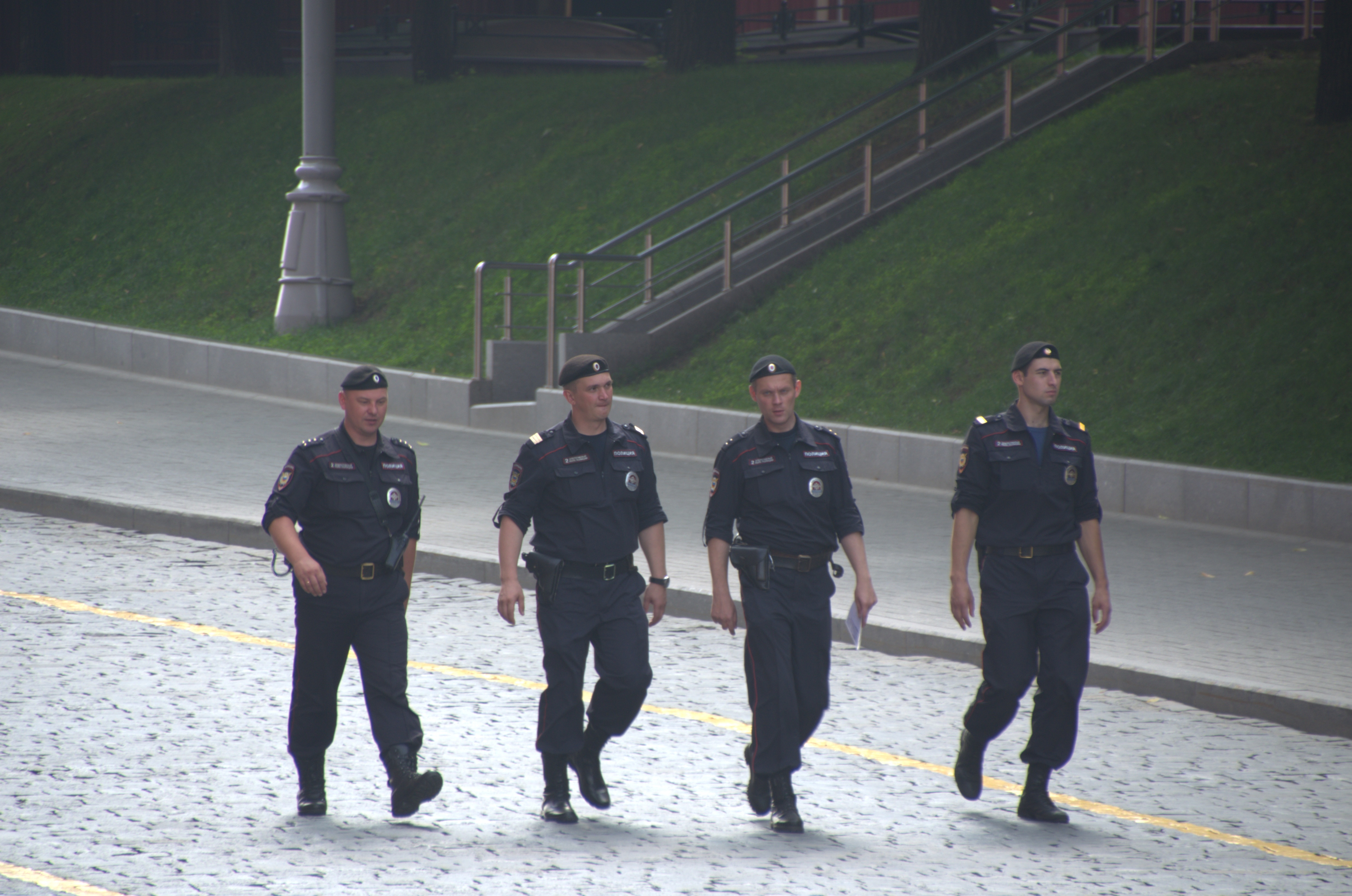 Vier Polizisten in Uniform gehen eine Straße entlang und halten Papiere in der Hand, mit einem Gebäude, Bäumen und einem Pfahl im Hintergrund.