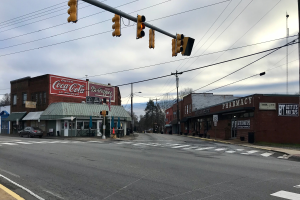 Eine Straßenkreuzung mit Ampel, Fahrzeugen, Gebäuden mit Schildern, Strommasten mit Kabeln, Bäumen und einem bewölkten Himmel.