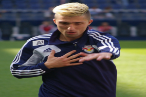 Ein junger Mann in einem blauen T-Shirt steht auf einem Fußballfeld und streckt die Arme aus, wobei er entschlossen als Flügelstürmer erscheint.