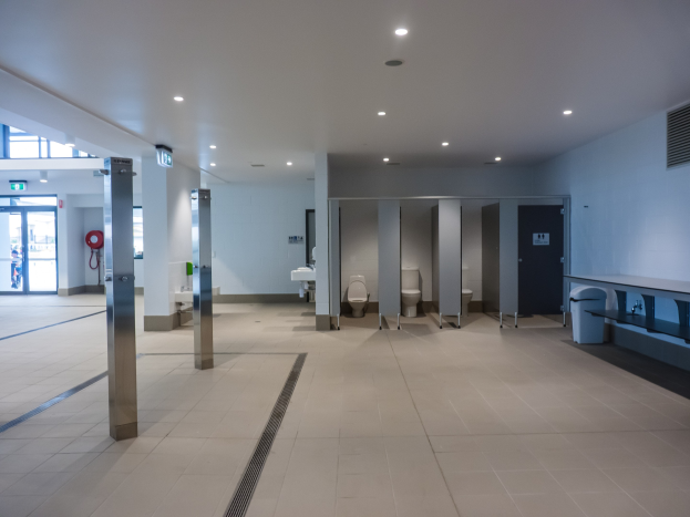 Großes öffentliches WC mit mehreren Toiletten, tragenden Säulen, einem Müllbehälter, einer Tür, einem Schild, einem Feuerlöscher und Deckenleuchten.