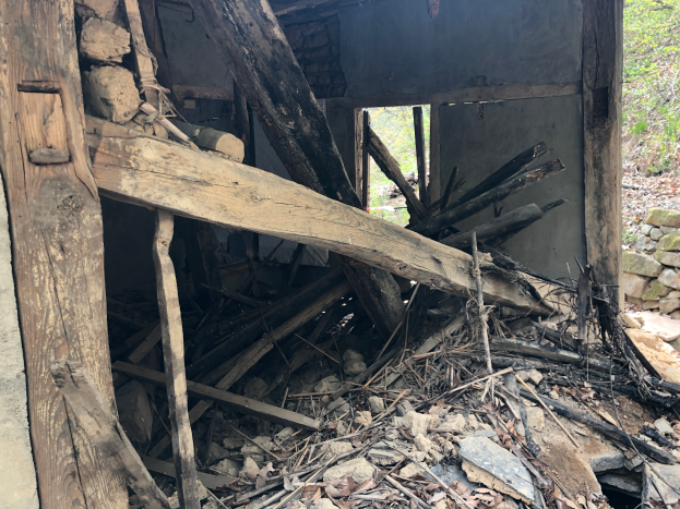 Ein altes Gebäude umgeben von Trümmer wie Holzstäben, Steinen und trockenen Blättern, mit Bäumen im Hintergrund, scheint im Abrissprozess zu sein.