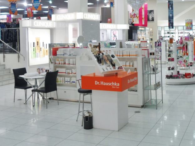 Ein mit verschiedenen Kosmetikartikeln gefüllter Laden auf Regalen, mit Stühlen, einem Tisch mit Gegenständen, einem Mülleimer, einem Kronleuchter, Schildern, einer Treppe mit Geländern, Kleidung an Bügeln und Deckenleuchten.