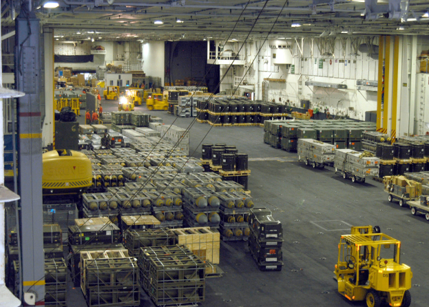 Ein großes Lagerinneres mit zahlreichen Trucks, Containern, Menschen, Lichtern, Stangen und einem prominenten Rad.