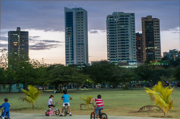 Kinder auf Fahrrädern in einem Park bei Sonnenuntergang, mit saftig grünem Gras und Bäumen, vor einer Kulisse aus hohen Wolkenkratzern, die sich vor einem orange-rosa-violetten Himmel abzeichnen.