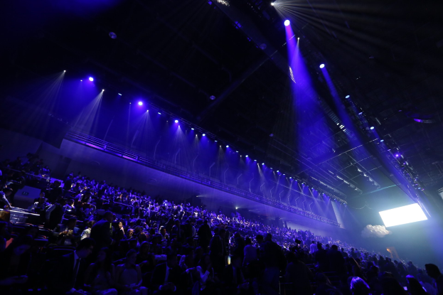 Eine große Menschenmenge sitzt vor einer Bühne mit Deckenbeleuchtung und einem Bildschirm auf der rechten Seite.