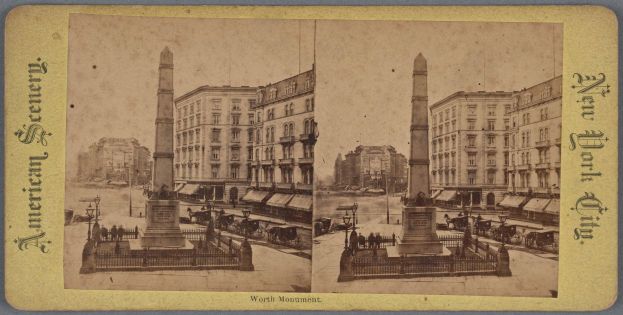Altes Schwarz-Weiß-Foto einer Stadtstraße mit einem zentralen Denkmal umgeben von Gebäuden, Straßenmöbeln, Fahrzeugen, Passanten und einem sichtbaren Himmel.