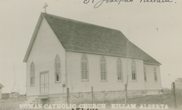 Ein Schwarz-Weiß-Foto von St. Joseph's lutherischer Kirche in Killam, Alberta, das ein Haus mit Dach, Fenstern, Türen und einem Kreuz auf dem Dach zeigt, umgeben von einem Zaun, Gras und anderen Häusern im Hintergrund, mit Text oben und unten im Bild.
