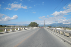 Eine Autobahn mit Motorradfahrern, umgeben von Geländern, Strommästen mit Käble, Bäumen, Hügeln und einem bewölkten Himmel.