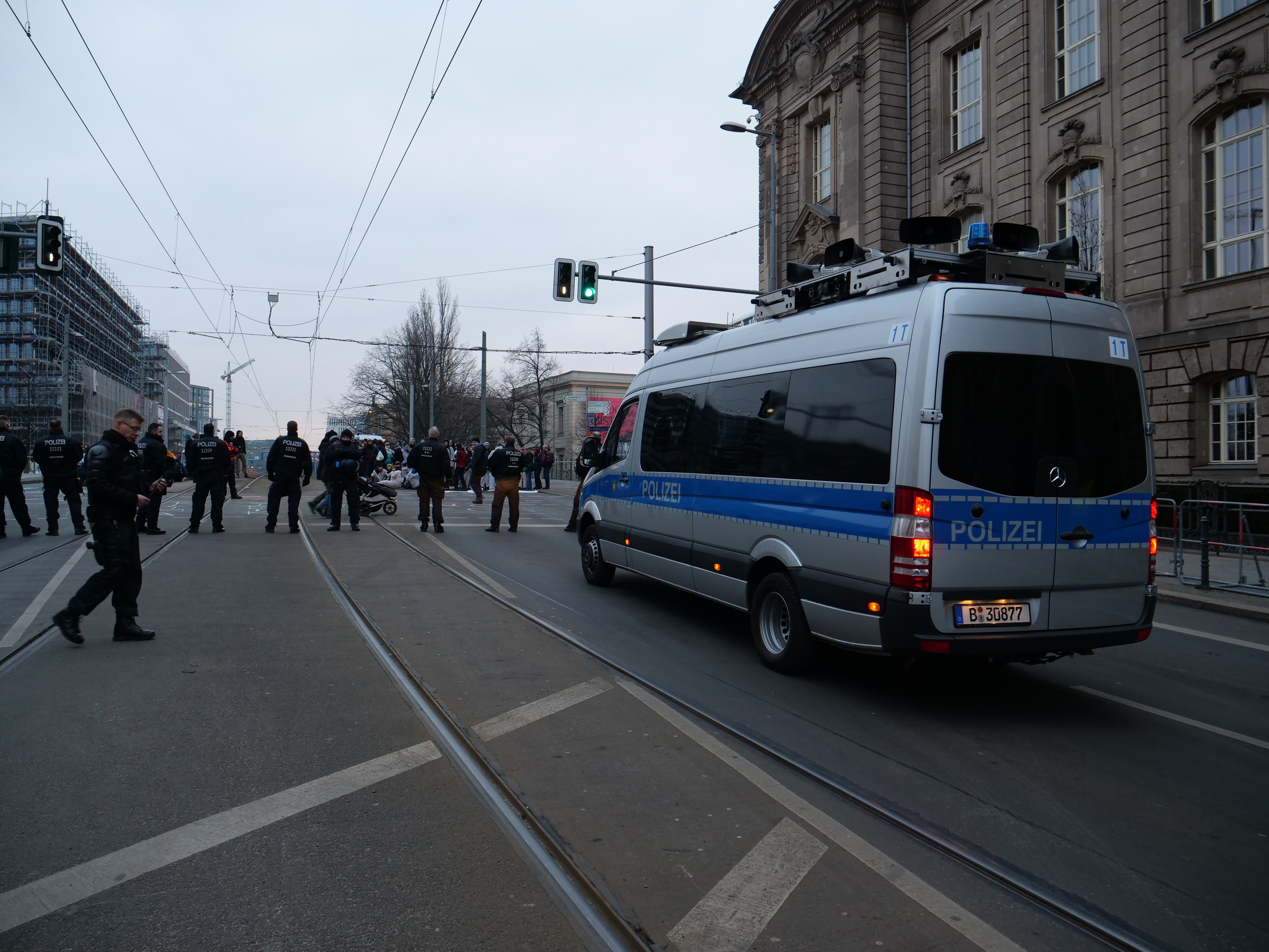 Ein Polizeifahrzeug steht an der Seite einer Straße, umgeben von einer Gruppe von Menschen in schwarzer Kleidung, mit Gebäuden, Bäumen, Pfählen, Verkehrszeichen, Drähten und einem klaren blauen Himmel im Hintergrund.