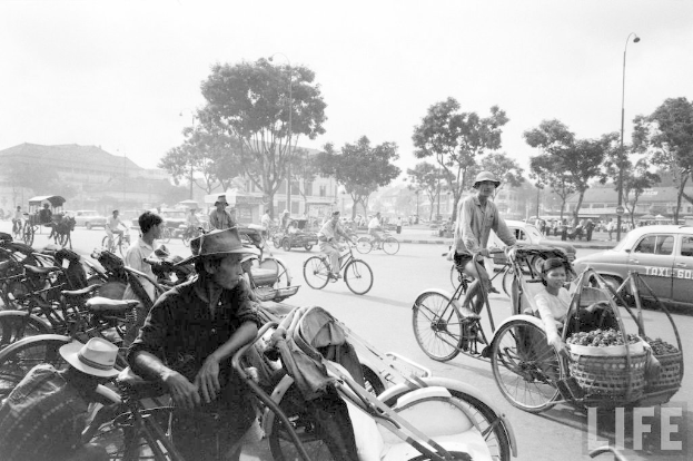 Eine vielbefahrene Straße mit Fahrrädern, Autos und anderen Fahrzeugen, mit Bäumen und Gebäuden im Hintergrund.