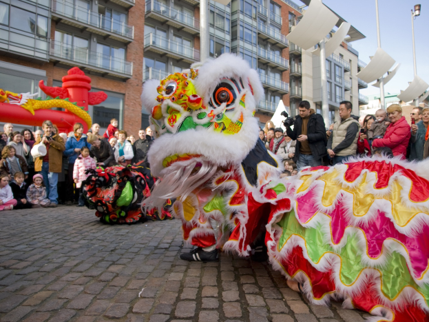 Ein lebendiges chinesisches Neujahrsfest in Amsterdam mit einem Löwen tanzen im Vordergrund und einer Menge Menschen drumherum, Gebäuden und Laternenmasten im Hintergrund und einem klaren blauen Himmel. Einige Menschen in der Menge halten Kameras.