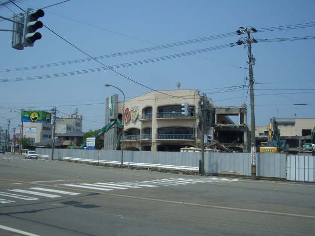 Stadtstraße mit Fahrzeugen, Strommästen, Laternen, einer Ampel auf der linken Seite, Gebäuden und Bäumen im Hintergrund und einem teilweise abgetragenen Gebäude im Vordergrund.