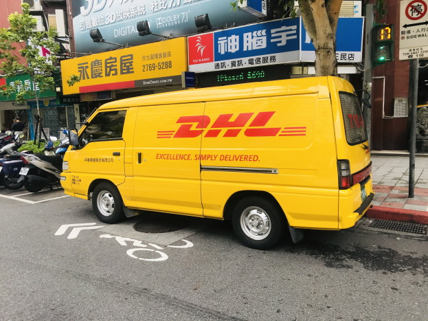 Gelbes DHL-Lieferfahrzeug auf Straße neben Gebäuden, Bäumen, Schildern, Masten, Ampeln und einer Person auf dem Gehweg.