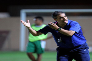 Ein Mann in einem blauen T-Shirt und Hose steht auf einem Fürballfeld und hält ein Mikrofon in der Hand, wobei er auf Spieler zeigt. Im Hintergrund ist eine Person in einem grünen Kleid und ein Torpfosten zu sehen.