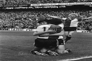 Drei Fußballspieler umarmen sich auf dem Boden und feiern ein Tor, mit einer jubelnden Menge und schwenkenden Fahnen im Hintergrund, dargestellt in Schwarz-Weiß.