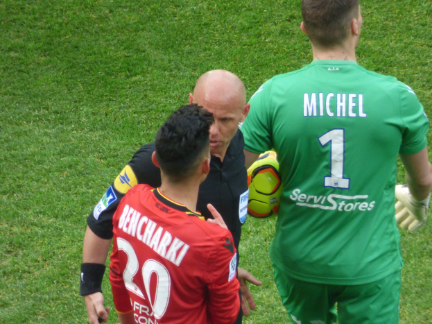 Schiedsrichter spricht mit zwei Füßballspielern auf einem saftig grünen Feld, ein Spieler hält einen Ball.