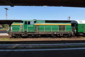 Ein grüner Diesel-Elektrolok auf Eisenbahnschienen unter einer Brücke, umgeben von Steinen, Gras, einem Pfad, Bäumen, Strommasten mit Drähten, einem Turm, Fahrzeugen und einem bewölkten Himmel.