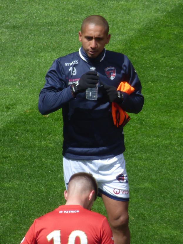 Ein Profisportler steht auf einem saftig grünen Feld, hält in einer Hand eine Flasche und in der anderen ein oranges Tuch.