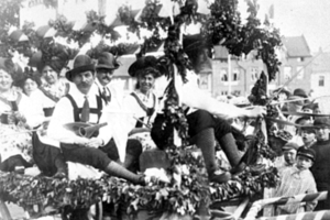 Schwarzes und weißes Bild eines Paradewagens mit Menschen in traditioneller bayrischer Tracht, einige spielen Instrumente, mit Blattdekorationen vor einem Hintergrund aus Gebäuden und einem klaren Himmel.