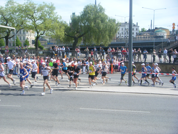 Gruppe von Läufern bei einem Marathon auf einer Straße mit Zuschauern, Absperrungen und städtischen Elementen unter einem bewölkten Himmel.