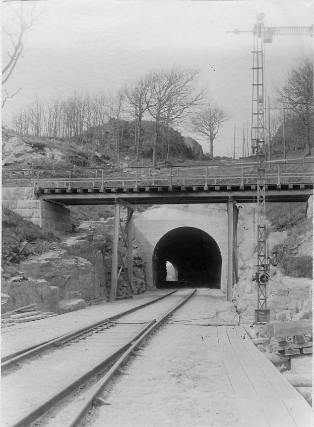Ein Schwarz-Weiß-Foto eines Tunnels im Bau mit Eisenbahnschienen, einer Brücke mit Pfeilern und Geländern, Bäumen im Hintergrund und einem Himmel darüber.
