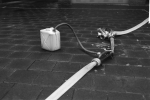 Schwarzes und weißes Foto eines Gasbehälters mit einem Schlauch, der vor einem Gebäude mit einer Tür im Hintergrund auf dem Boden steht.