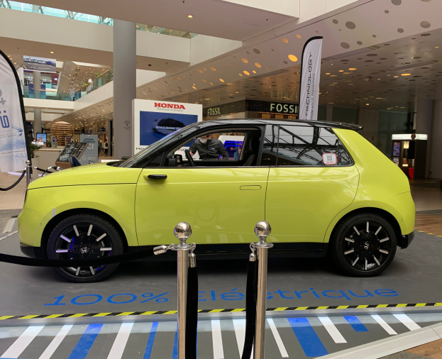 Ein gelbes wasserstoffbetriebenes Elektroauto vor einem Gebäude geparkt, umgeben von Absperrpfosten mit Seilen, Bannern und Schildern, mit einer Person im Auto, einer Gruppe von Menschen in der Nähe, einer Säule, einem Topf mit einer Pflanze, Deckenleuchten und einer Decke mit Deckenventilatoren.