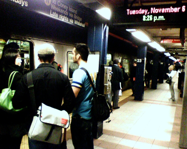 Eine volle Zugplatform mit Menschen, die Taschen tragen, ein Zug auf der linken Seite und ein Textanzeigebrett oben.